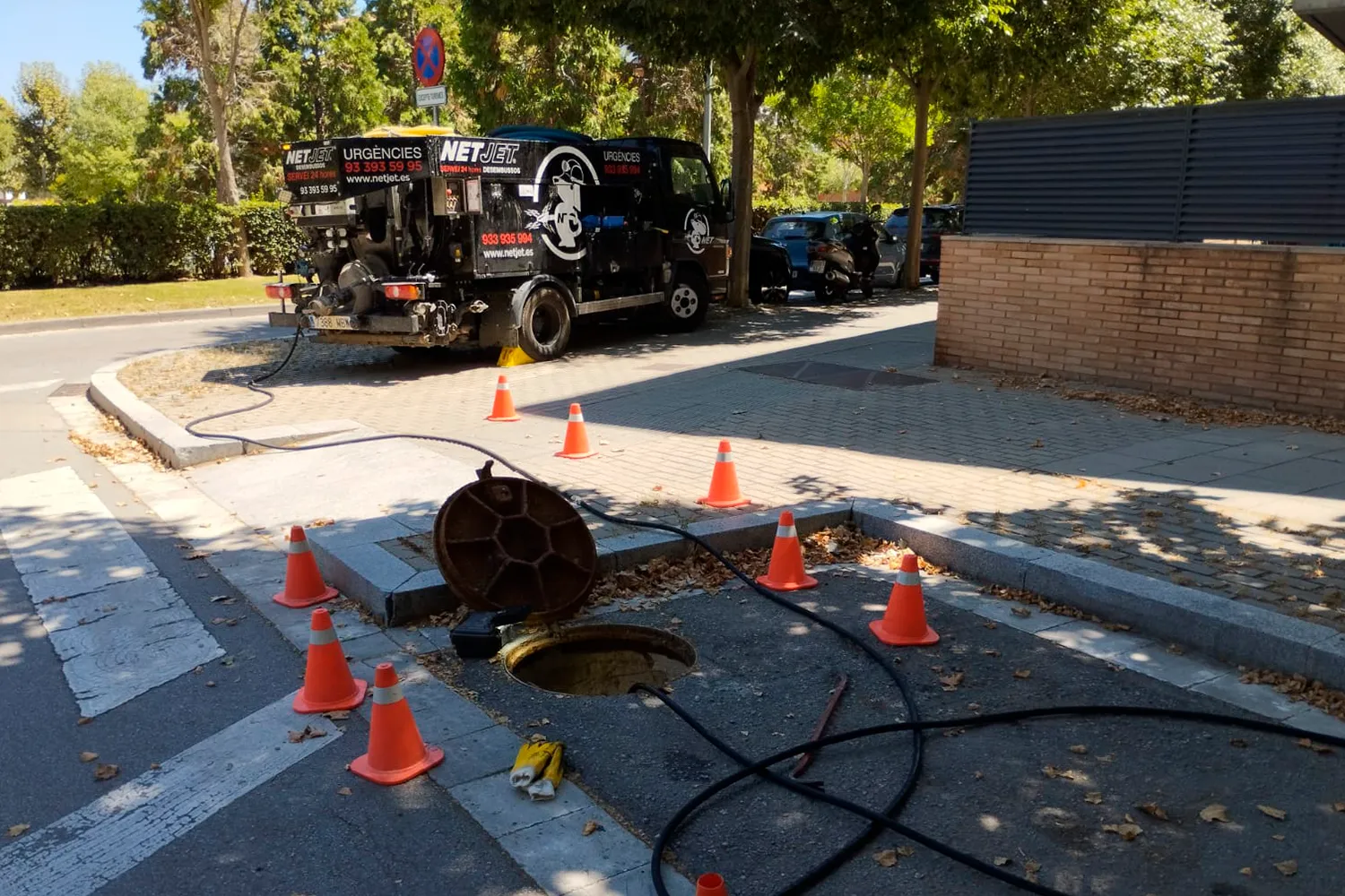 La imagen muestra una escena al aire libre, donde se está llevando a cabo una inspección o limpieza de alcantarillado. En primer plano, se puede ver una tapa de alcantarilla abierta rodeada por varios conos de tráfico de color naranja, que sirven para delimitar la zona de trabajo y advertir a los transeúntes y vehículos. Dos mangueras largas y negras salen de la alcantarilla y se conectan a un camión que está estacionado en la acera.