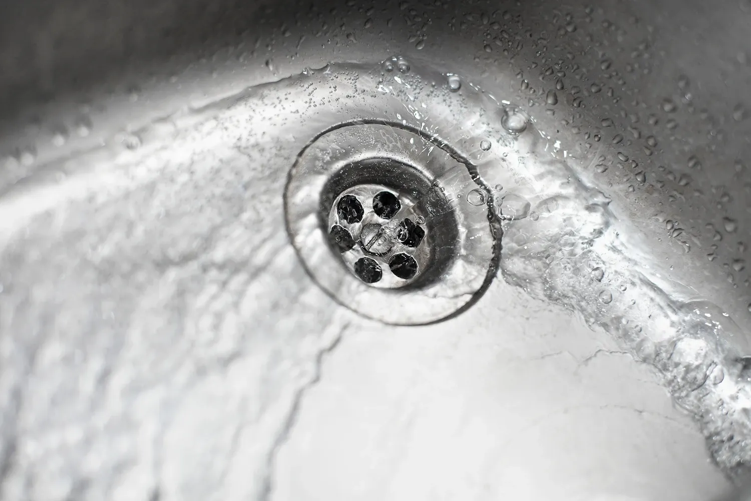 La imagen muestra un desagüe de un fregadero, con agua fluyendo hacia el drenaje. El fregadero parece estar hecho de acero inoxidable y tiene gotas de agua dispersas por su superficie, lo que sugiere que el agua ha estado corriendo o salpicando recientemente. El desagüe tiene una rejilla metálica, que permite que el agua pase mientras retiene objetos más grandes. La escena resalta la importancia de un drenaje limpio y funcional, crucial en cocinas y baños, para evitar atascos y asegurar un flujo adecuado de agua.