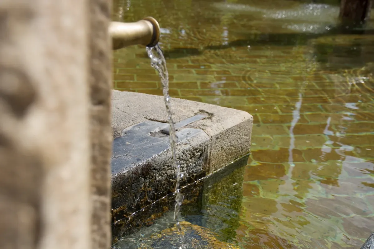 a imagen muestra un primer plano de una fuente de agua, con agua fluyendo de un grifo hacia una pila de piedra abajo. El agua es clara y refleja la luz circundante, creando un efecto centelleante en la superficie. La pila de piedra parece ser parte de una fuente o característica de agua más grande, con detalles adicionales desenfocados en el fondo.