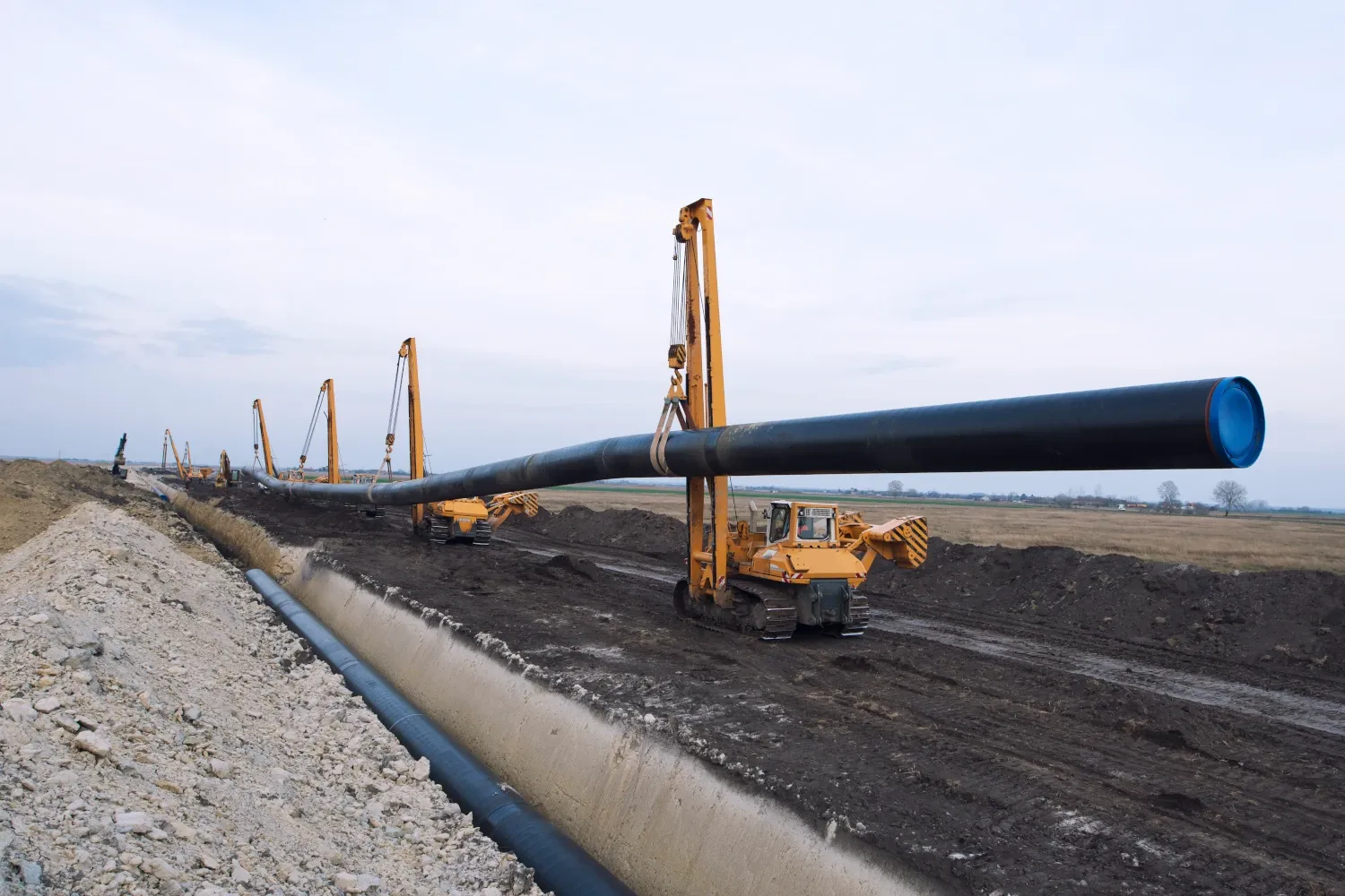 La imagen muestra un proceso de instalación de una tubería de gran tamaño en un entorno abierto, probablemente en una zona rural o industrial. Varias grúas móviles, de color amarillo, están alineadas a lo largo del trazado de la tubería, levantando y posicionando secciones de la misma sobre una zanja que parece destinada a enterrarla. La escena refleja un proyecto de construcción de infraestructura, posiblemente un gasoducto o acueducto, con maquinaria especializada trabajando en el levantamiento y colocación precisa de las tuberías sobre el terreno excavado. El cielo está despejado, y el entorno es mayormente llano.