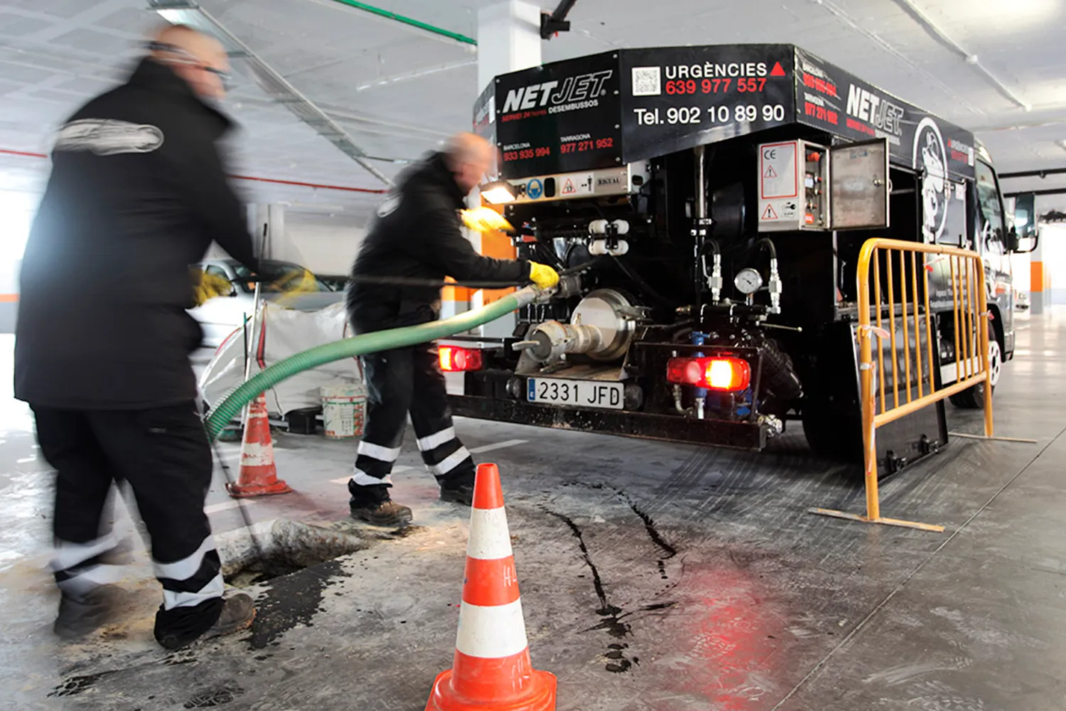 La imagen muestra a dos operarios de la empresa "NetJet" realizando tareas de mantenimiento en un estacionamiento subterráneo. Están usando equipo especializado conectado a un camión de la empresa, que está equipado con tecnología para la gestión de aguas residuales. Los trabajadores están manipulando una manguera verde que parece estar insertada en una alcantarilla o pozo de inspección en el suelo.