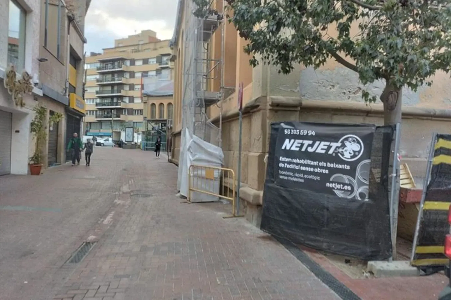 La imagen muestra una calle tranquila en una zona urbana, donde se están llevando a cabo trabajos de rehabilitación en un edificio. A la derecha, junto al edificio, se observa una lona negra con el logotipo y la información de la empresa "NetJet", especializada en servicios de inspección y rehabilitación de bajantes. El texto en la lona indica que la empresa ofrece servicios sin necesidad de obra, destacando su enfoque económico, rápido, ecológico y con mínimas molestias.