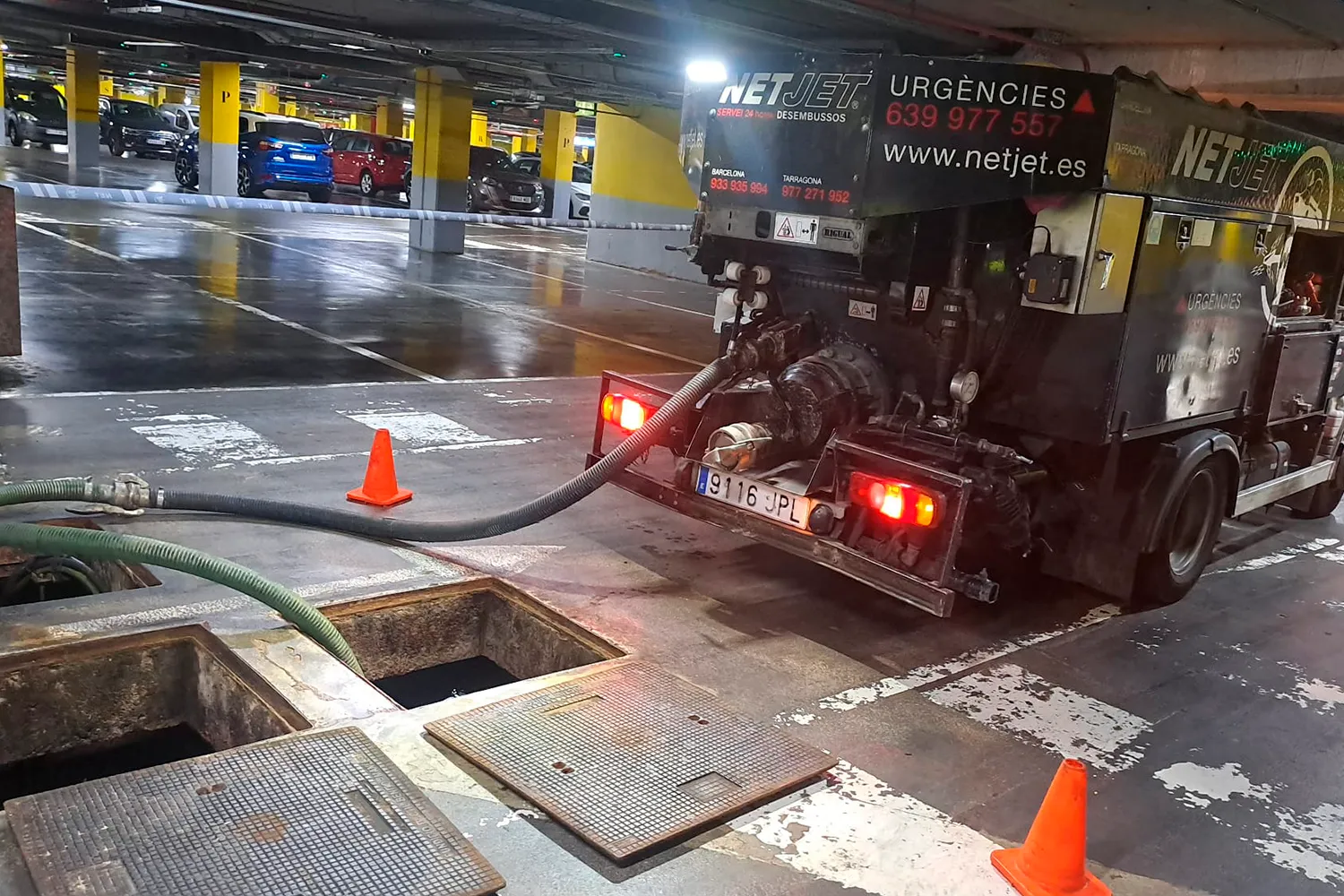 La imagen muestra una escena en un estacionamiento subterráneo, con un camión de servicios de limpieza de alcantarillado en primer plano. El camión, de la empresa "NetJet", tiene varios letreros visibles, como el nombre de la empresa, un número de teléfono para emergencias y su sitio web. El vehículo está conectado a una alcantarilla abierta a través de mangueras, probablemente para realizar trabajos de limpieza o desatasco. Hay un par de conos de color naranja colocados alrededor del área de trabajo para señalar la intervención.