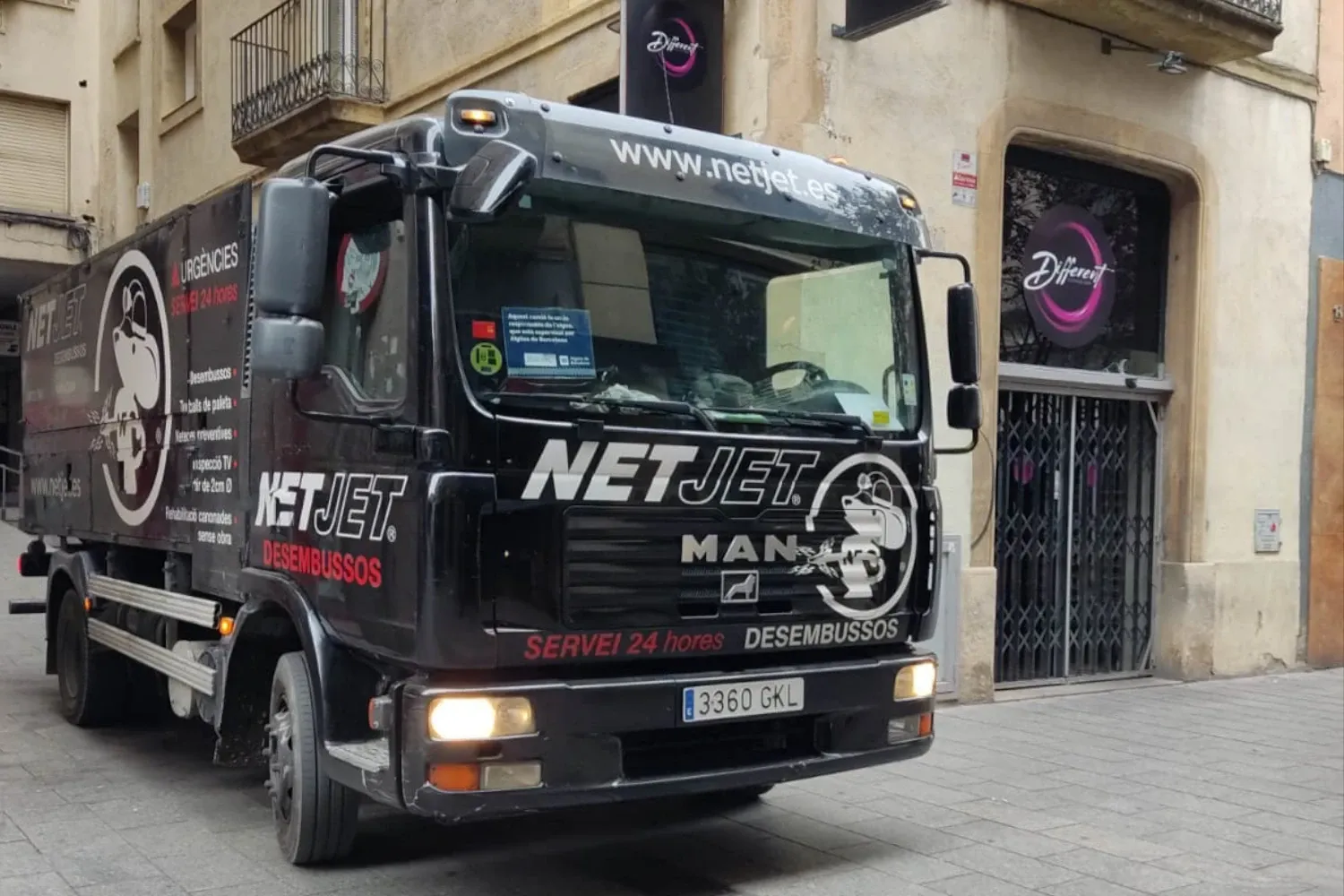 La imagen muestra un camión de la empresa NetJet estacionado en una calle urbana, frente a un edificio con una fachada clásica. 
Este tipo de vehículo está diseñado para manejar emergencias relacionadas con el alcantarillado, y la presencia de la rotulación "Servei 24 hores" indica que NetJet está disponible las 24 horas del día para atender situaciones urgentes. La imagen enfatiza la especialización y el compromiso de NetJet en ofrecer soluciones rápidas y efectivas para el mantenimiento de infraestructuras urbanas.